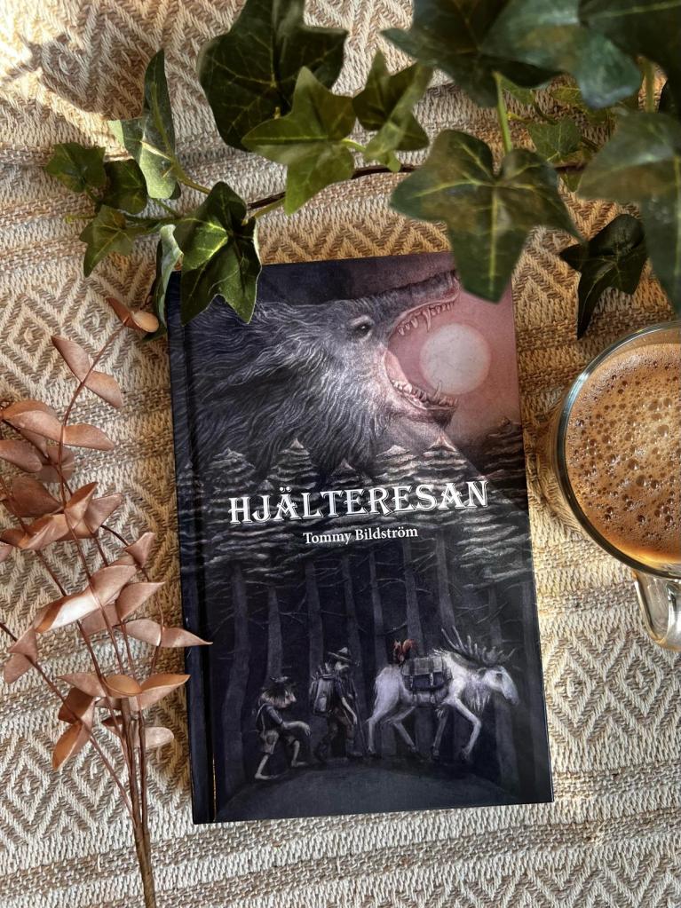 Boken Hjälteresan av Tommy Bildström ligger på en duk omgiven av en kaffekopp och ett par kvistar. På bokslaget syns en illustration av en varg som sväljer en sol och en skog där en älg och två människoliknande varelser vandrar.