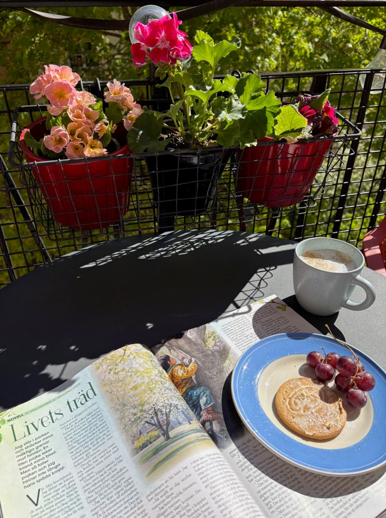 Min novell Livets träd syns i en uppslagen tidning på ett balkongbord tillsammans med fika och blommande krukväxter i bakgrunden.